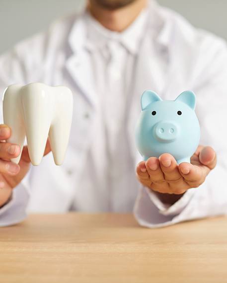 Dentist in white jacket holding a model tooth and blue piggy bank