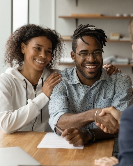 Smiling man shaking hands with person across desk next to smiling woman